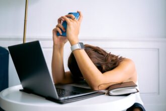 Profissional demonstrando extrema exaustão mental ao descansar a cabeça sobre uma mesa de trabalho, segurando um smartphone azul. A pessoa usa smartwatch e está cercada por equipamentos de trabalho (laptop, óculos, caderno), representando visualmente os sintomas de burnout e sobrecarga digital. A postura corporal expressa esgotamento profissional e fadiga causada pelo excesso de conexão tecnológica e pressão no ambiente corporativo.