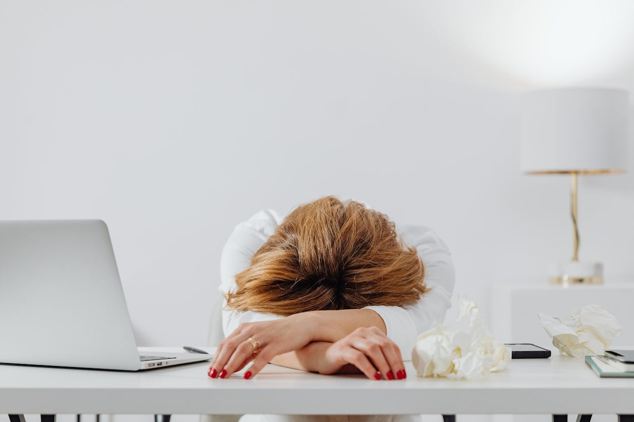 Mulher com cabelos castanhos presos em coque, vestindo blusa branca, descansando a cabeça sobre os braços cruzados em uma mesa de trabalho branca. A cena retrata esgotamento profissional em um ambiente de escritório clean e moderno, com laptop aberto, smartphone, flores brancas decorativas e uma luminária dourada ao fundo. A postura corporal expressa claramente os sintomas de fadiga mental e burnout no ambiente corporativo contemporâneo.