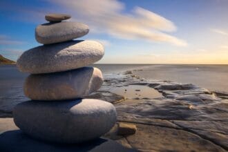 Torre de pedras empilhadas em equilíbrio perfeito sobre formação rochosa natural, com vista para o mar ao pôr do sol. A imagem simboliza a harmonia e concentração necessárias para como meditar efetivamente, representando os pilares fundamentais da prática meditativa: equilíbrio mental, serenidade interior e conexão com a natureza. O cenário transmite a essência de como meditar em um ambiente tranquilo e inspirador.