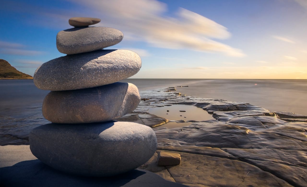 Torre de pedras empilhadas em equilíbrio perfeito sobre formação rochosa natural, com vista para o mar ao pôr do sol. A imagem simboliza a harmonia e concentração necessárias para como meditar efetivamente, representando os pilares fundamentais da prática meditativa: equilíbrio mental, serenidade interior e conexão com a natureza. O cenário transmite a essência de como meditar em um ambiente tranquilo e inspirador.