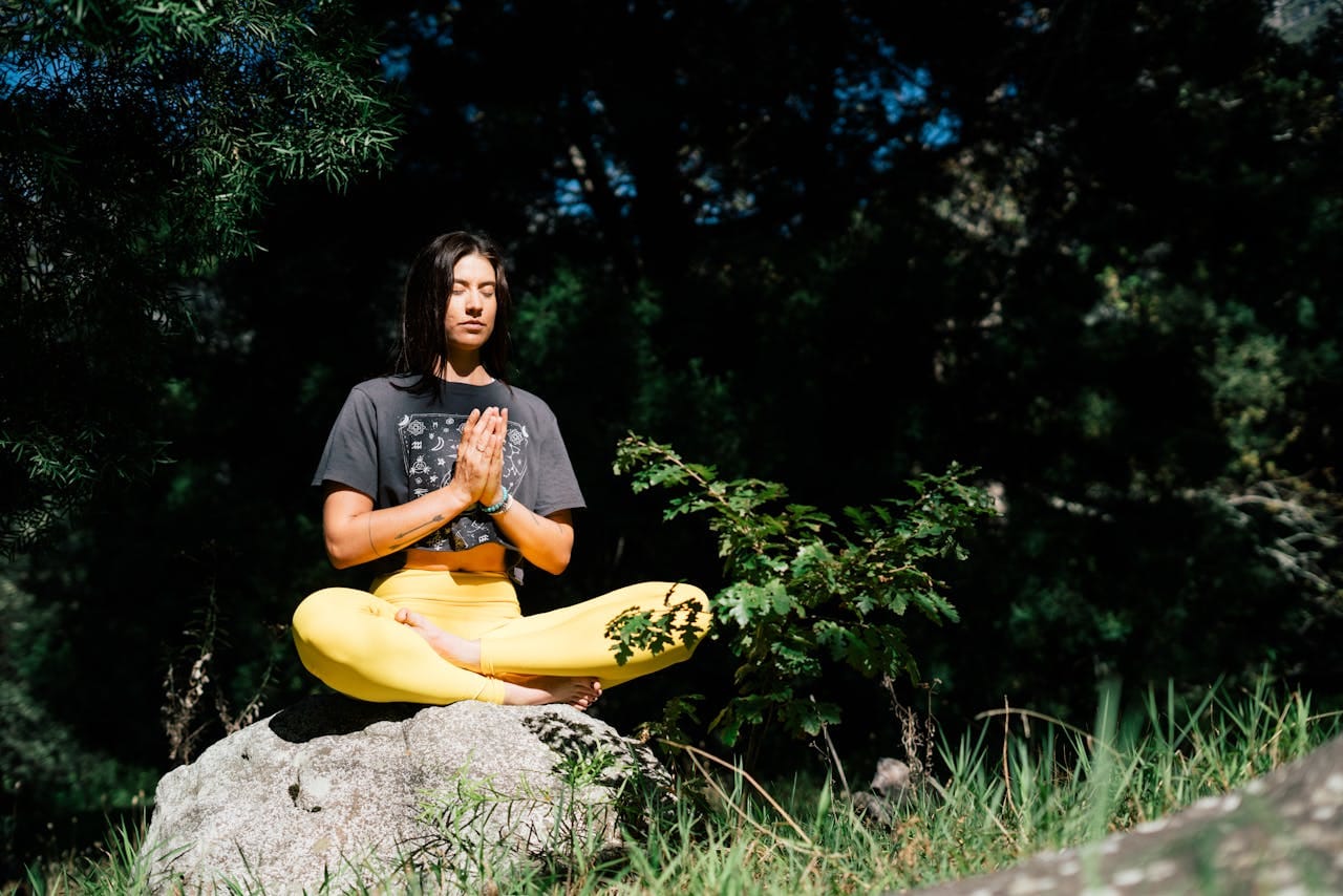 Mulher demonstrando como meditar na posição de lótus, sentada sobre uma pedra em ambiente natural cercado por vegetação exuberante. Vestindo camiseta cinza e calça amarela, ela mantém as mãos em posição de mudra (oração) com olhos fechados em concentração. A cena ilustra perfeitamente a técnica de como meditar ao ar livre, conectando-se com a natureza para alcançar paz interior e mindfulness através da prática meditativa tradicional