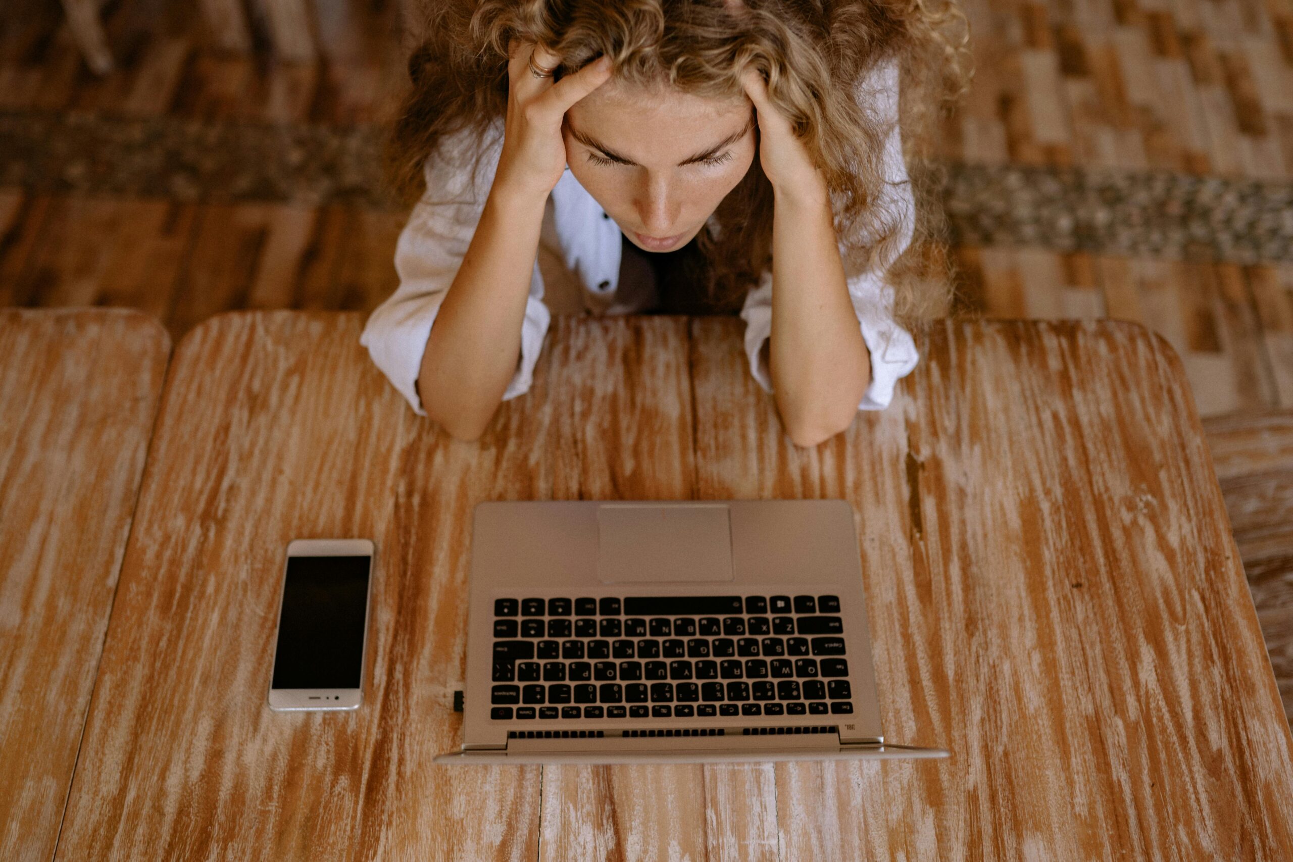 Mulher jovem com cabelos cacheados loiros demonstrando sinais de estresse e esgotamento profissional. Ela está sentada em frente a um laptop, com as mãos segurando a cabeça em uma postura que expressa fadiga mental e sobrecarga de trabalho. A cena retrata o ambiente de trabalho moderno, com smartphone ao lado, ilustrando perfeitamente os sintomas visuais da síndrome de burnout no contexto profissional atual