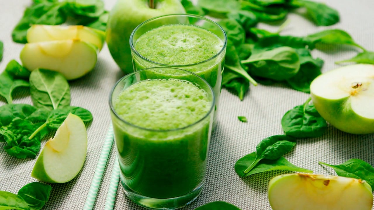 Suco verde detox natural servido em copos de vidro, preparado com ingredientes frescos como maçã verde, espinafre e folhas verdes variadas. A bebida detox apresenta textura cremosa e cor verde vibrante, ideal para desintoxicar o organismo. Composição saudável sobre fundo claro mostra os ingredientes naturais utilizados no preparo do chá verde detox, incluindo vegetais folhosos e frutas frescas que auxiliam na limpeza do corpo e eliminação de toxinas