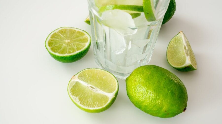 Água detox com limão fresco servida em copo de vidro, preparada com rodelas de limão e gelo para desintoxicar o organismo naturalmente. A bebida detox apresenta limões frescos cortados ao redor do copo sobre fundo branco minimalista. Esta água aromatizada com limão é um dos melhores chás e bebidas naturais para limpeza do organismo, auxiliando na eliminação de toxinas, acelerando o metabolismo e promovendo hidratação saudável. Receita simples e refrescante de água detox cítrica ideal para desintoxicação diária.