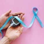 Mãos segurando glicosímetro para medição de diabetes com laço azul de conscientização sobre fundo rosa. A imagem simboliza o controle glicêmico e a importância de desintoxicar o organismo para quem tem diabetes. O medidor de glicose representa o monitoramento da saúde metabólica, enquanto o laço azul turquesa destaca a conscientização sobre diabetes e saúde preventiva. Chás detox e hábitos saudáveis ajudam a equilibrar os níveis de açúcar no sangue e promovem a desintoxicação natural do organismo em pessoas diabéticas ou pré-diabéticas.