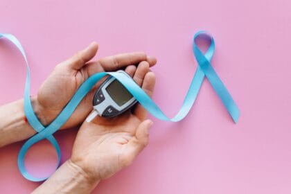 Mãos segurando glicosímetro para medição de diabetes com laço azul de conscientização sobre fundo rosa. A imagem simboliza o controle glicêmico e a importância de desintoxicar o organismo para quem tem diabetes. O medidor de glicose representa o monitoramento da saúde metabólica, enquanto o laço azul turquesa destaca a conscientização sobre diabetes e saúde preventiva. Chás detox e hábitos saudáveis ajudam a equilibrar os níveis de açúcar no sangue e promovem a desintoxicação natural do organismo em pessoas diabéticas ou pré-diabéticas.