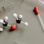 Modelo molecular científico com equipamentos de laboratório representando a composição química das toxinas no organismo e processos de desintoxicação. A estrutura molecular ao lado de proveta graduada e pipeta ilustra como substâncias químicas e compostos tóxicos se acumulam no corpo. Esta representação científica demonstra a importância de desintoxicar o organismo através de chás detox que auxiliam na eliminação de metais pesados, radicais livres e outras toxinas em nível celular. A química da desintoxicação natural envolve compostos bioativos presentes em chás e ervas que promovem a limpeza profunda do fígado e rins.