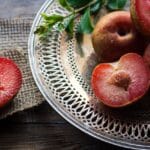Ameixas vermelhas maduras inteiras e cortadas ao meio dispostas em prato branco vazado com folhas verdes sobre toalha de juta e mesa de madeira envelhecida